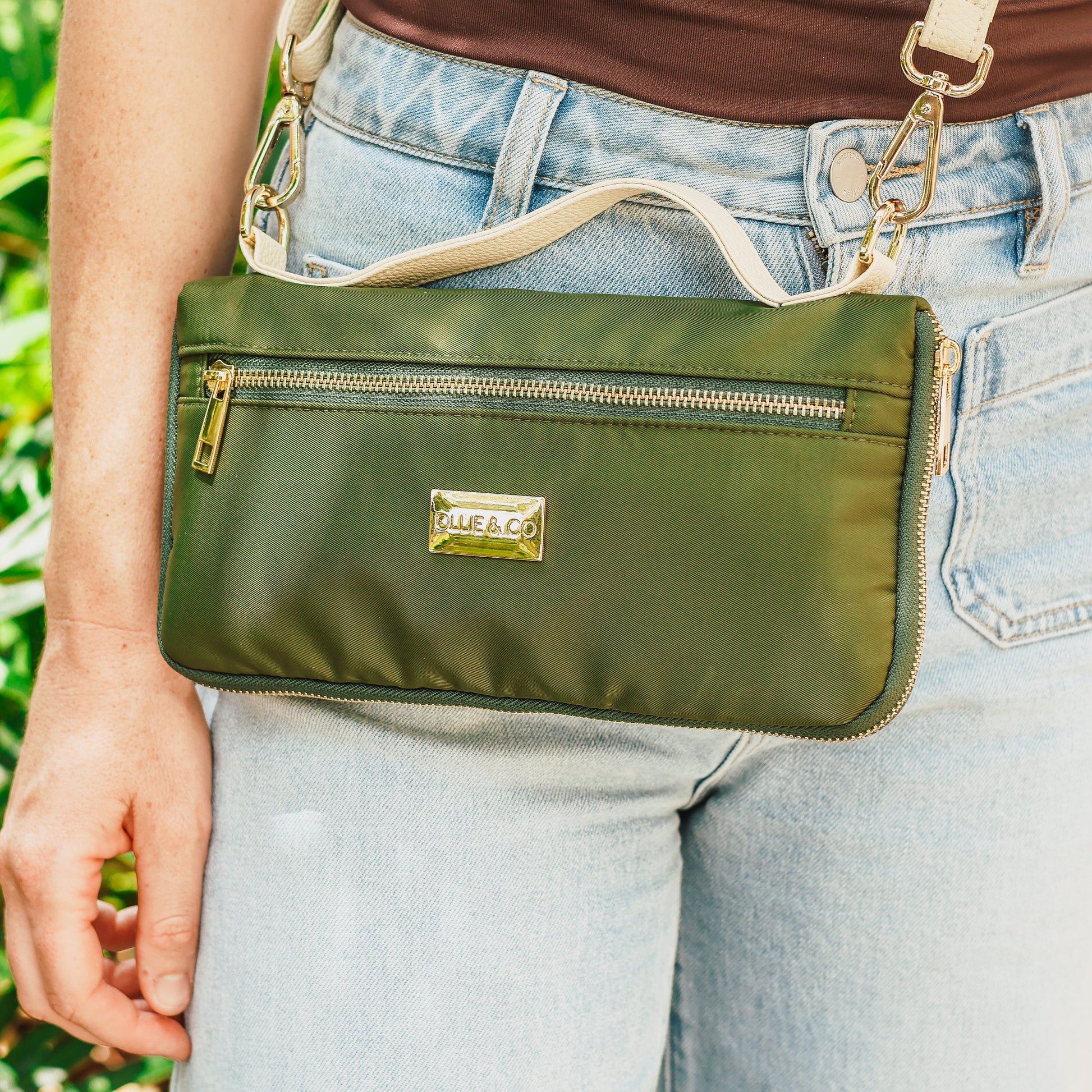 Green handbag with gold zipper and brand logo held by a person wearing jeans.